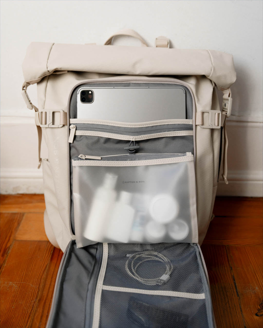 A beige and gray backpack shown open, with a tablet inserted into the top compartment and toiletry bottles and a cable visible in the front mesh pockets. The bag is photographed indoors on a wooden floor against a white wall.