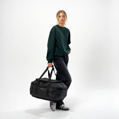 A woman stands in a studio wearing a dark green sweatshirt, dark jeans, and sneakers while carrying a medium black duffel bag.