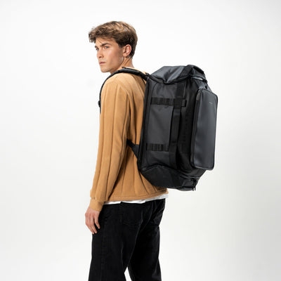 A man wearing a tan sweater and black pants is shown from the side/back profile carrying a medium black backpack against a clean white studio background.