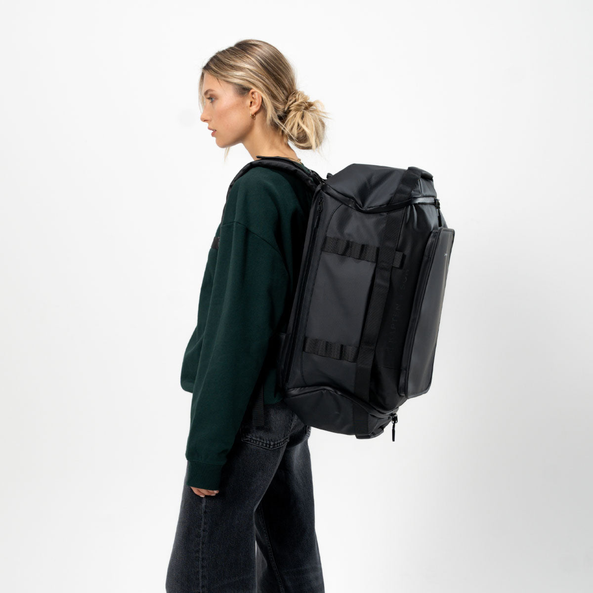 A woman in a dark green sweatshirt and dark jeans is shown from the side, wearing a medium sized black duffel backpack with multiple compression straps and a top opening.