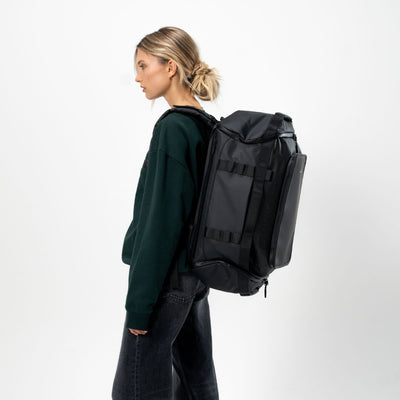 A woman in a dark green sweatshirt and dark jeans is shown from the side, wearing a medium sized black duffel backpack with multiple compression straps and a top opening.