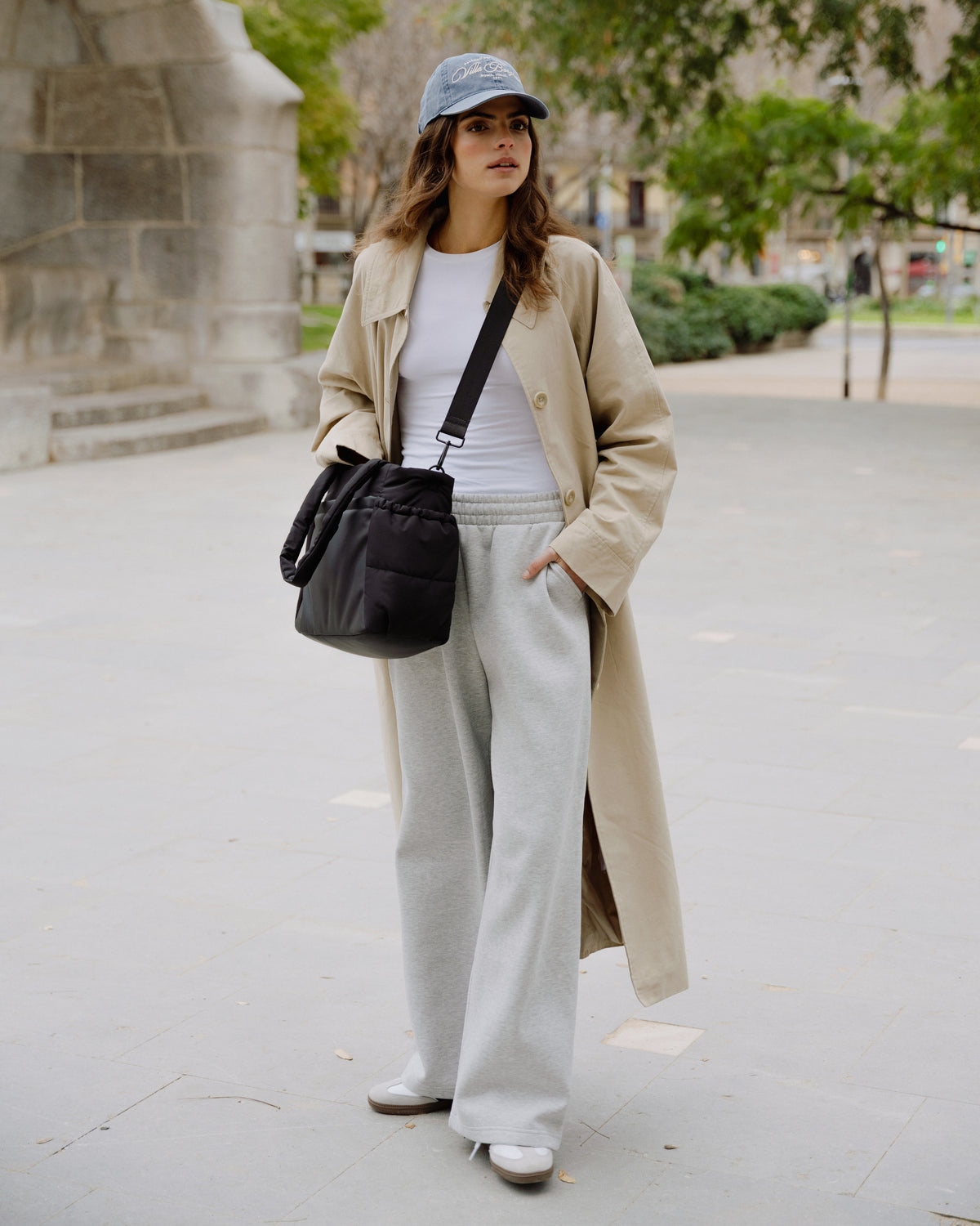 A woman stands outdoors on a paved walkway wearing a long beige trench coat over a white top and light gray wide-leg sweatpants, with a black crossbody bag and a blue cap.