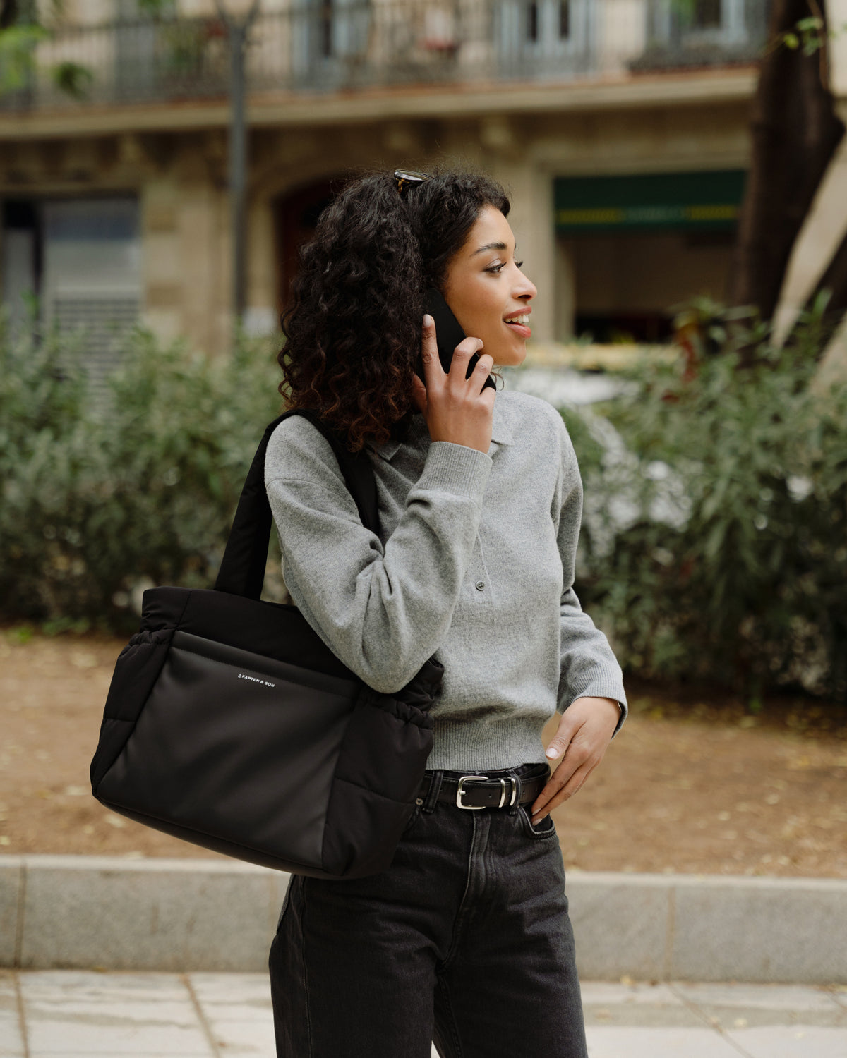 A woman with curly dark hair stands outdoors on a sidewalk, holding a phone to her ear while wearing a black shoulder bag across her body. She is dressed in a gray knit sweater, black jeans, and a black belt, with a blurred urban background of buildings and greenery.