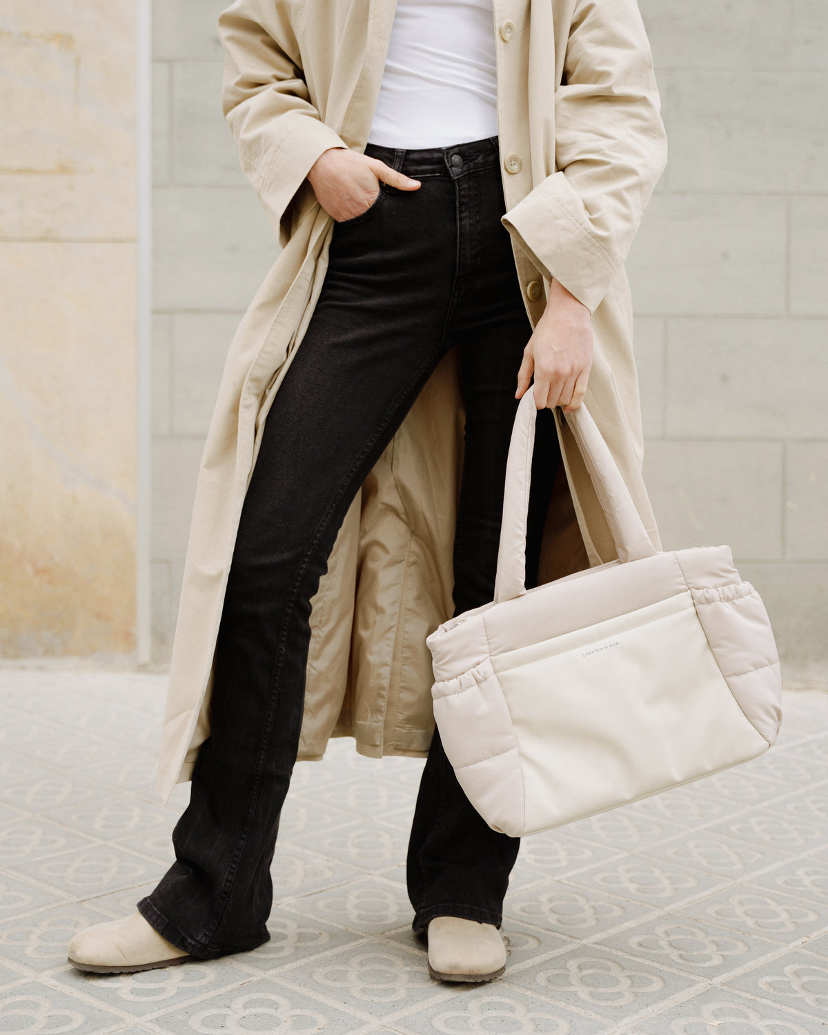 A person wearing a long beige trench coat over a white top and black flared jeans, holding a large cream quilted tote bag. The scene is set against a light stone wall and patterned tile floor.