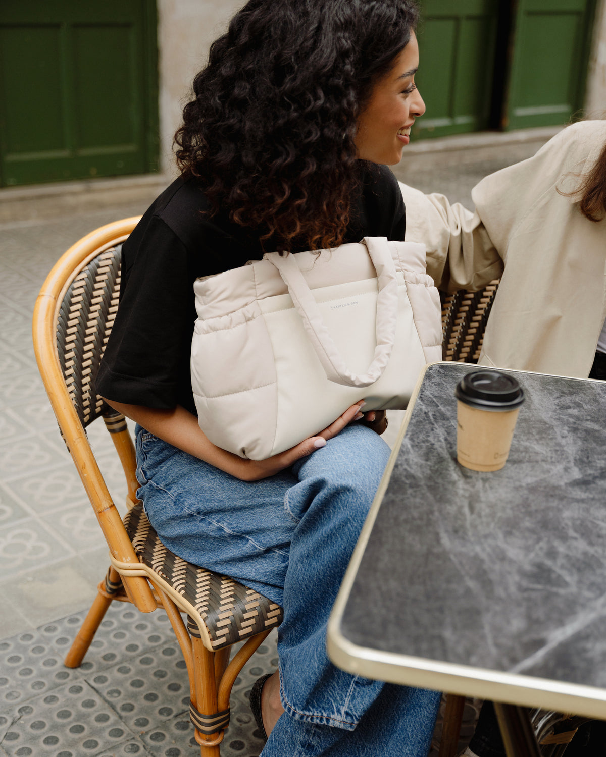 A woman sitting at an outdoor cafe table holding a large cream quilted puffer tote bag. The scene includes a woven chair, a coffee cup on the table, and another person partially visible at the table.