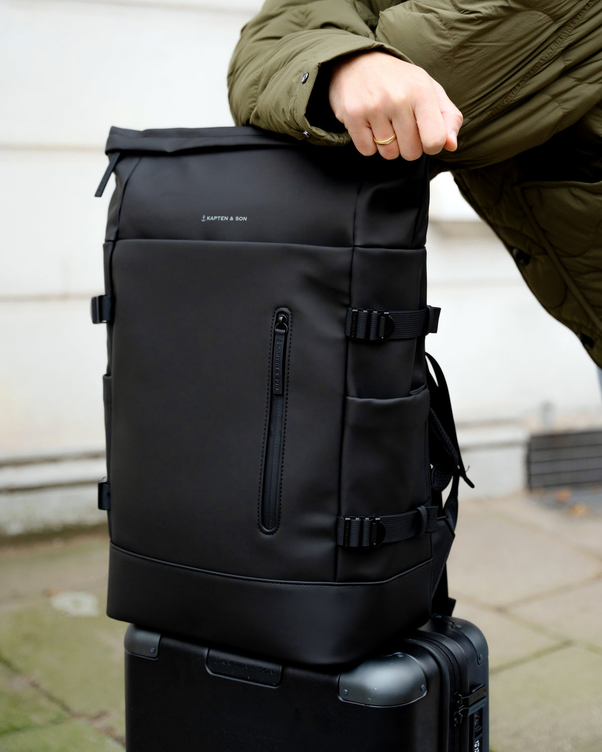 A black backpack with multiple zippered compartments and buckle straps, shown outdoors with a person's hand resting on top and a suitcase underneath.
