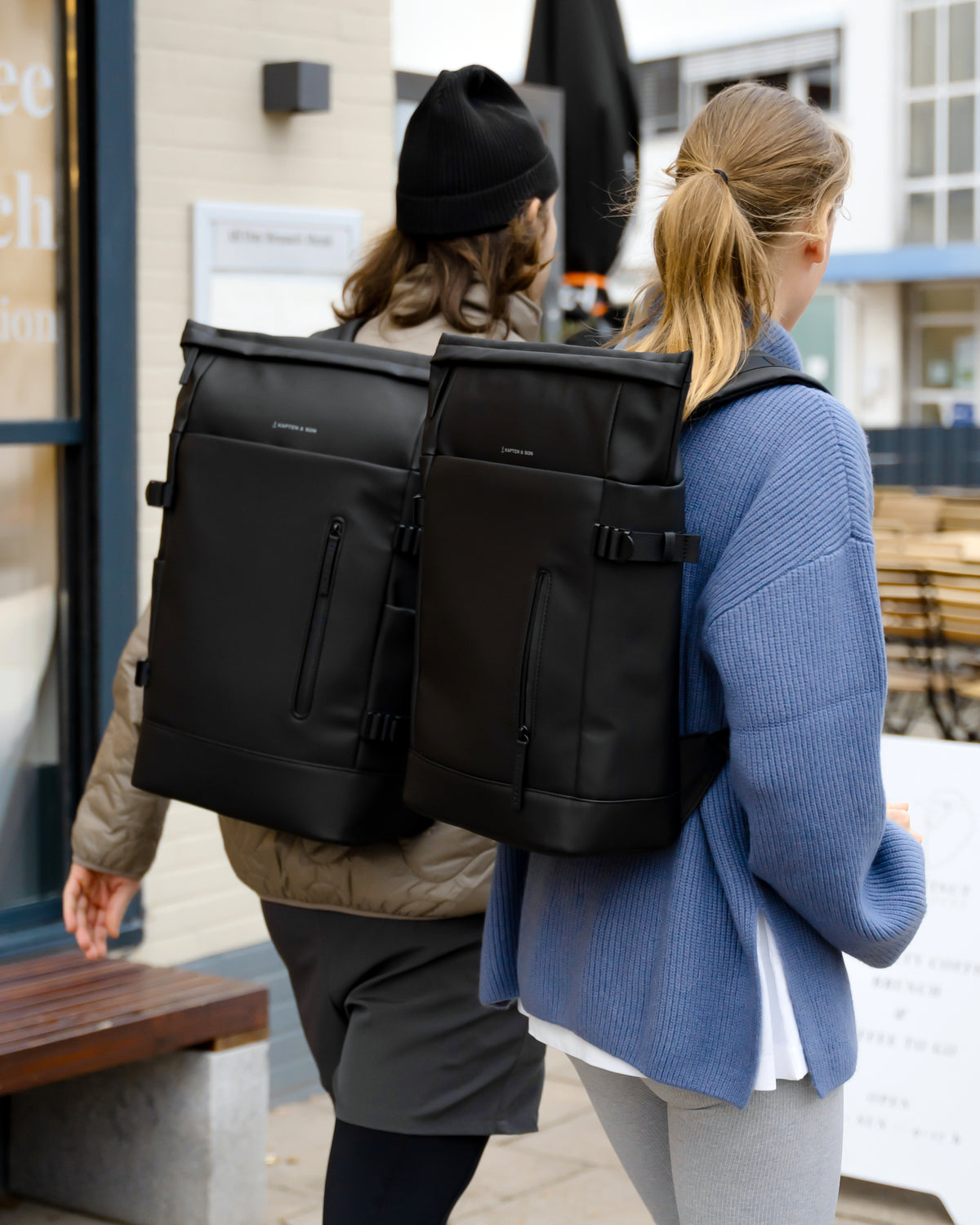 Two women walking away from the camera, each wearing a large black dual-compartment backpack designed to carry two laptops. The backpacks feature a matte finish, zippered compartments, and side compression straps.