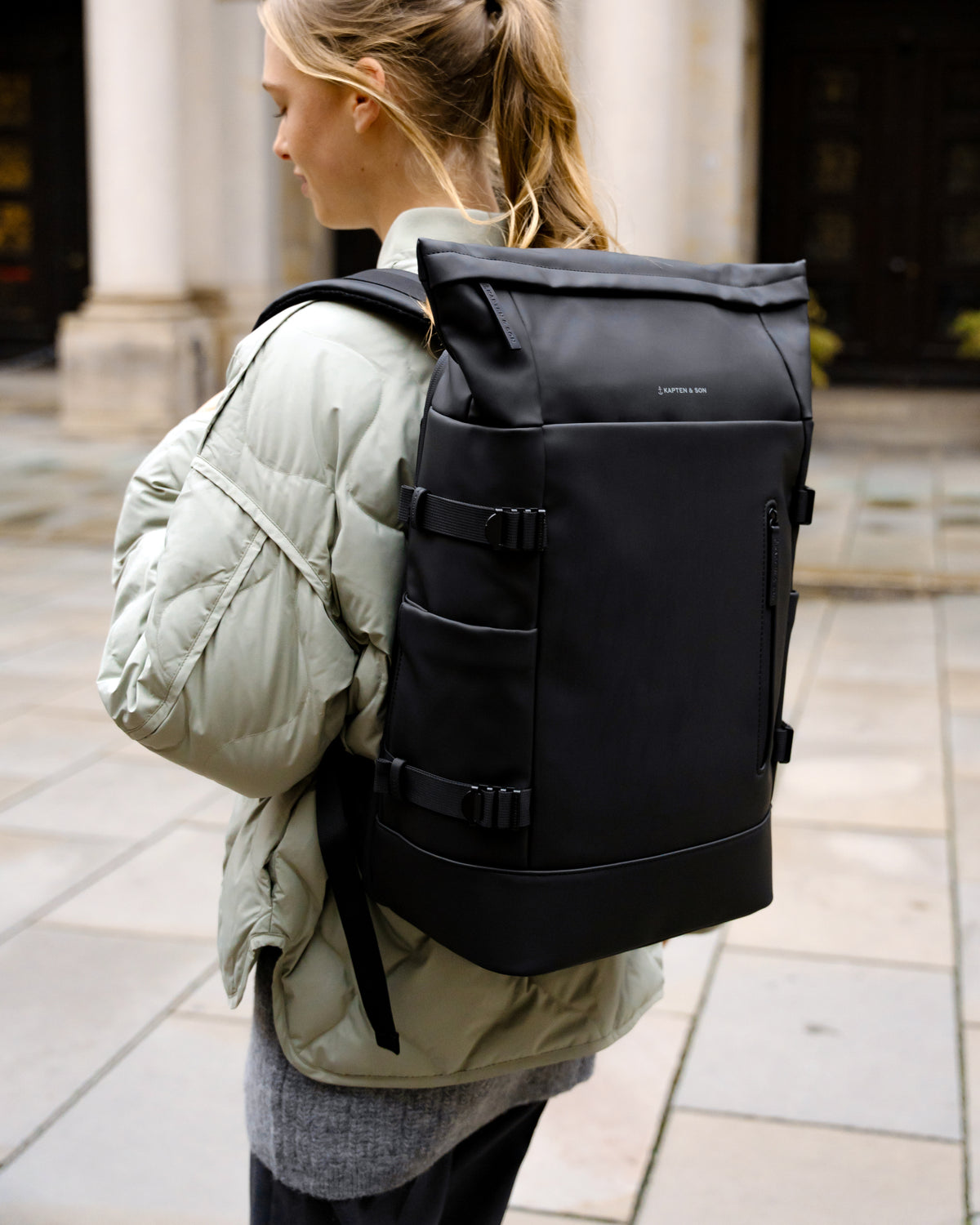 A woman wearing a light olive green puffer jacket carries a black water resistant backpack with multiple straps and a top flap, walking outdoors on a stone paved walkway.