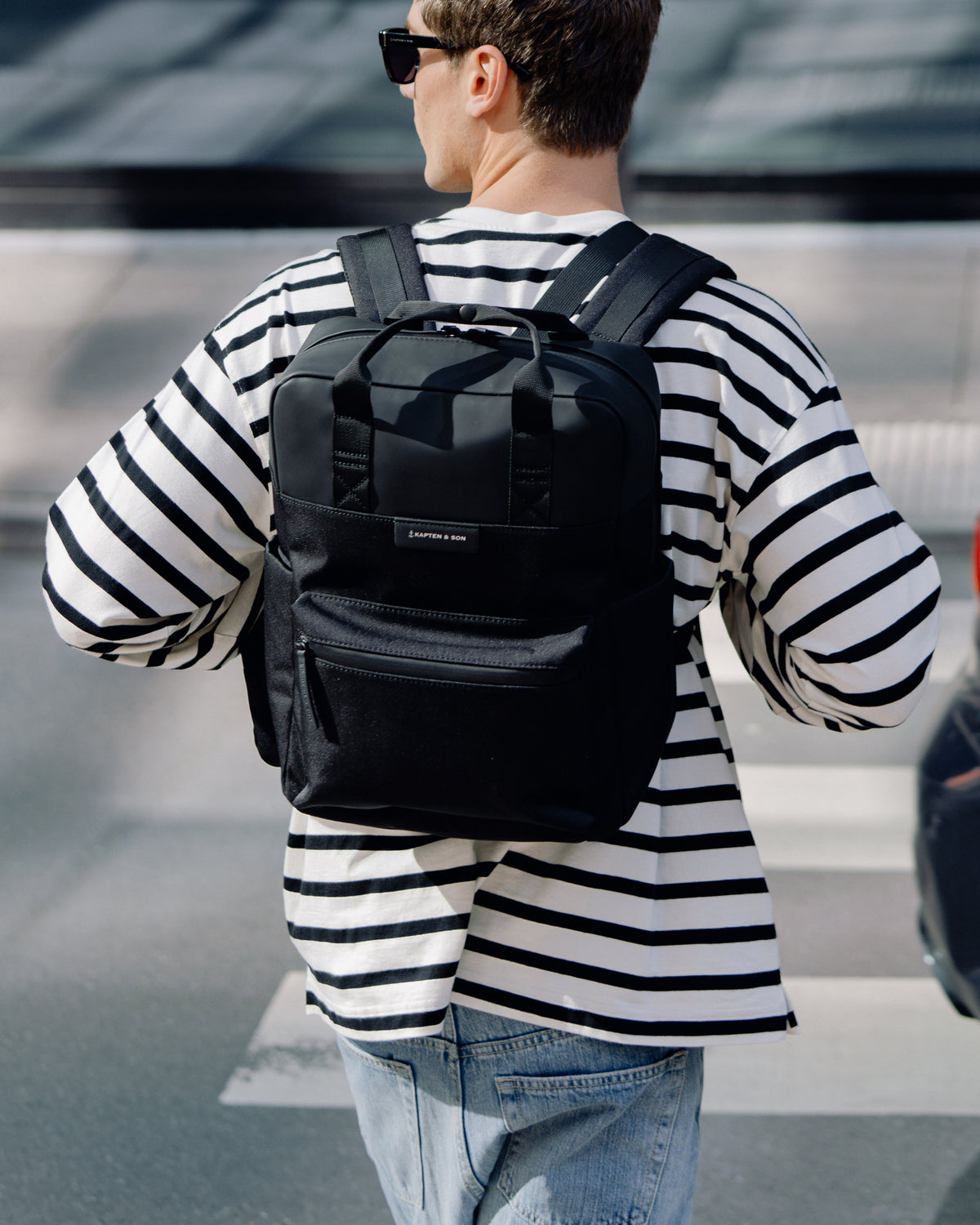 A man wearing a black backpack with a front zippered pocket and top carry handles, styled with a black and white striped long-sleeve shirt and light blue jeans.