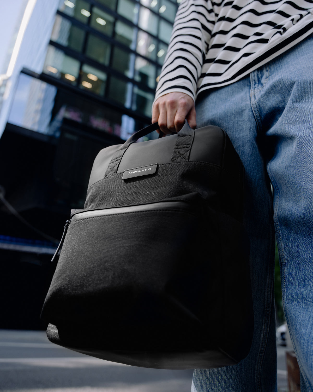 A person in a black and white striped shirt and blue jeans carries a black fabric backpack with a top handle and front zip pocket in an outdoor urban setting.
