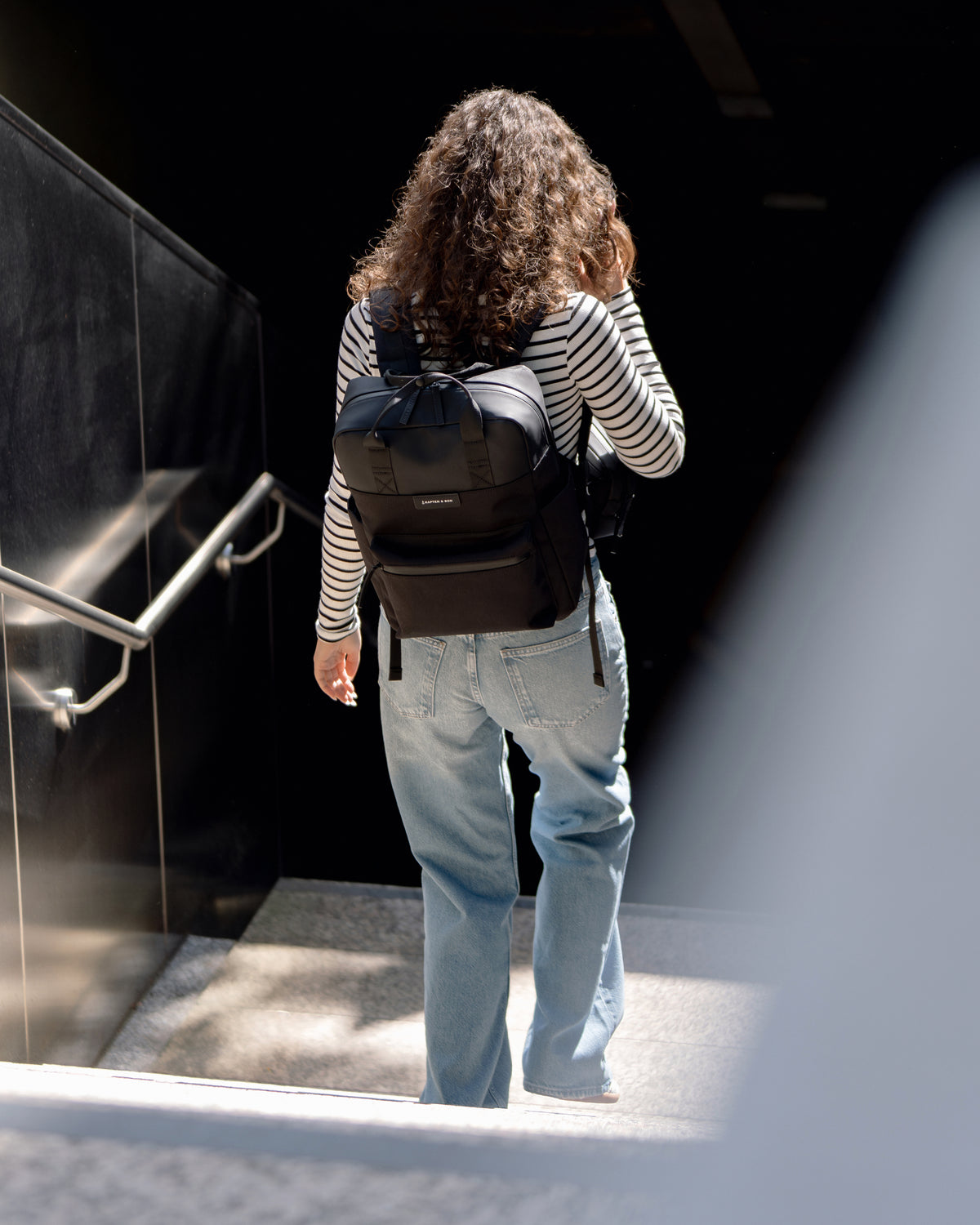 A woman with curly hair seen from behind walking down a staircase, wearing a black backpack, a black and white striped long-sleeve shirt, and light blue jeans.