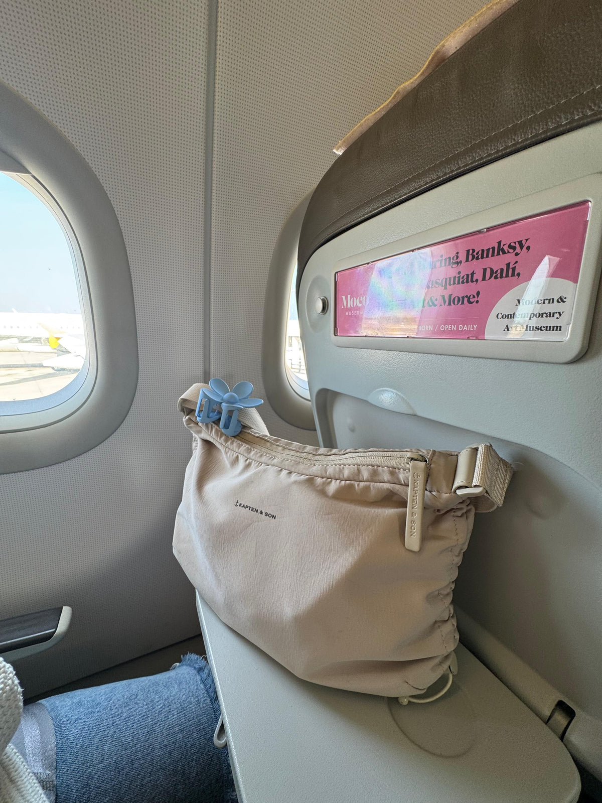 A beige shoulder bag with a blue hair clip accent resting on an airplane seat next to the window.
