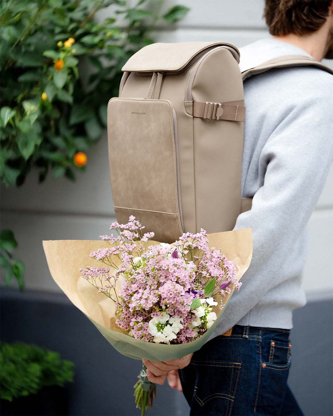 A person wearing a light gray sweatshirt and blue jeans carries a taupe beige backpack while holding a bouquet of pink and white flowers wrapped in brown paper. The photo is taken outdoors with green foliage and a white wall in the background.