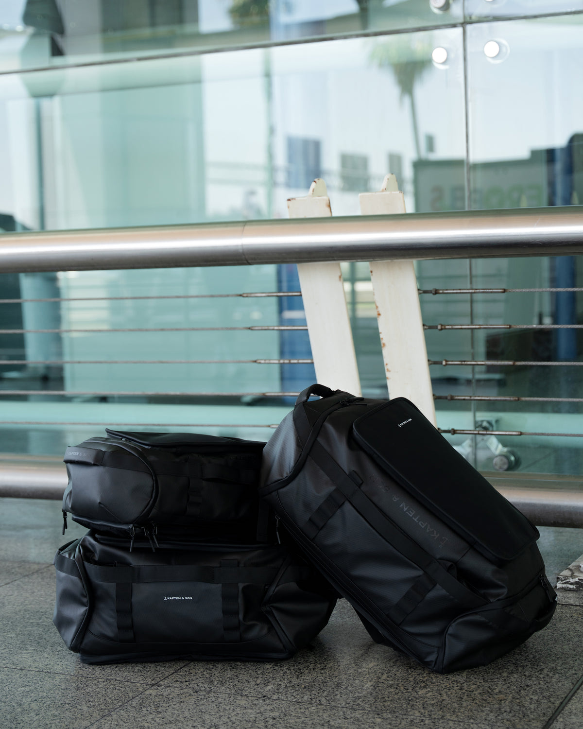 Three black duffel bags stacked against a glass wall with metal railing.