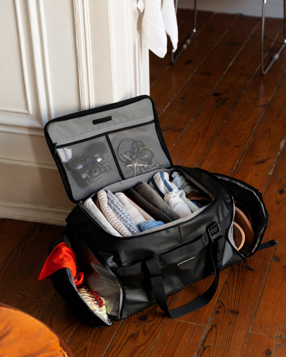 A black travel duffel bag with multiple compartments, shown open on a wooden floor next to a white wall. The bag is filled with folded clothes and accessories, including a red garment in the side pocket, a white towel hanging in the background, and a metal chair leg visible in the upper right.