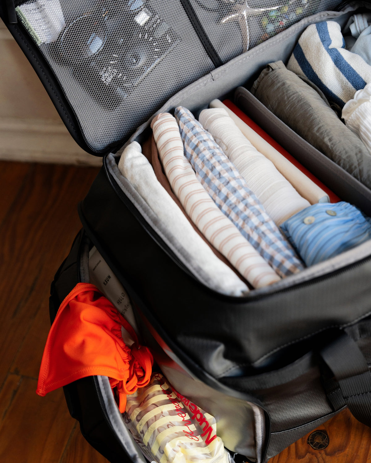 An open black packing bag filled with neatly folded clothes and toiletries, photographed on a wooden floor in a real indoor setting.