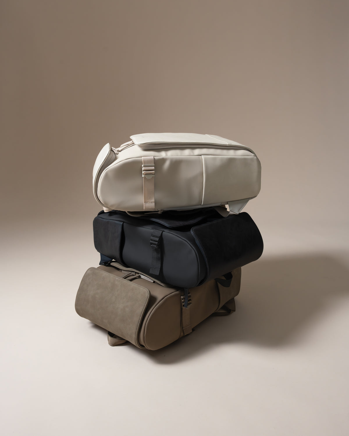 Three duffel bags stacked vertically on a warm beige studio background. The top bag is a light cream/off-white color, the middle is black, and the bottom is taupe-brown.
