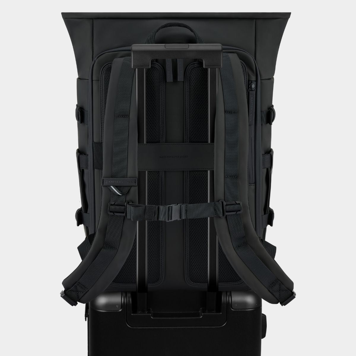 The back view of a black rolling suitcase with a large black backpack mounted on top, set against a light gray studio background.