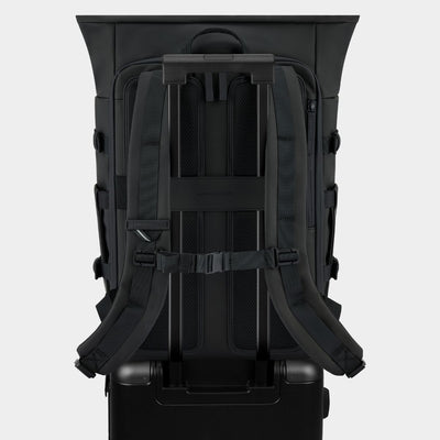 The back view of a black rolling suitcase with a large black backpack mounted on top, set against a light gray studio background.
