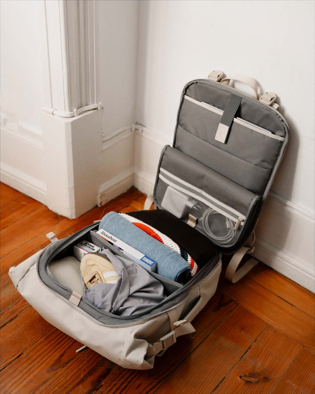 An open gray and beige backpack resting on a wooden floor against a white wall, with folded clothing and accessories visible inside.