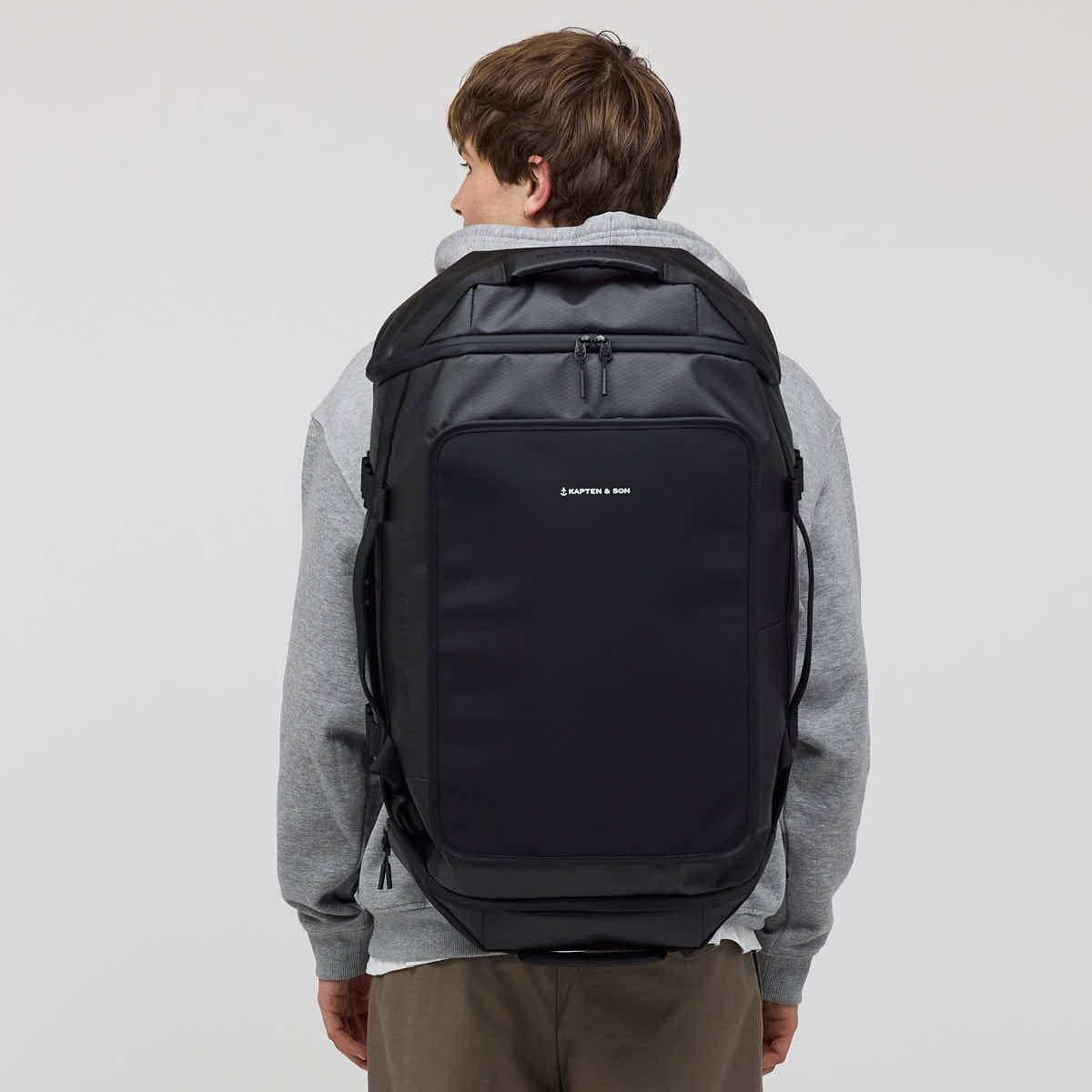A man viewed from behind wearing a black backpack with a gray hoodie and brown pants, set against a plain light gray studio background. The backpack features a front zippered pocket with a small white 'KAPTEN & SON' logo.