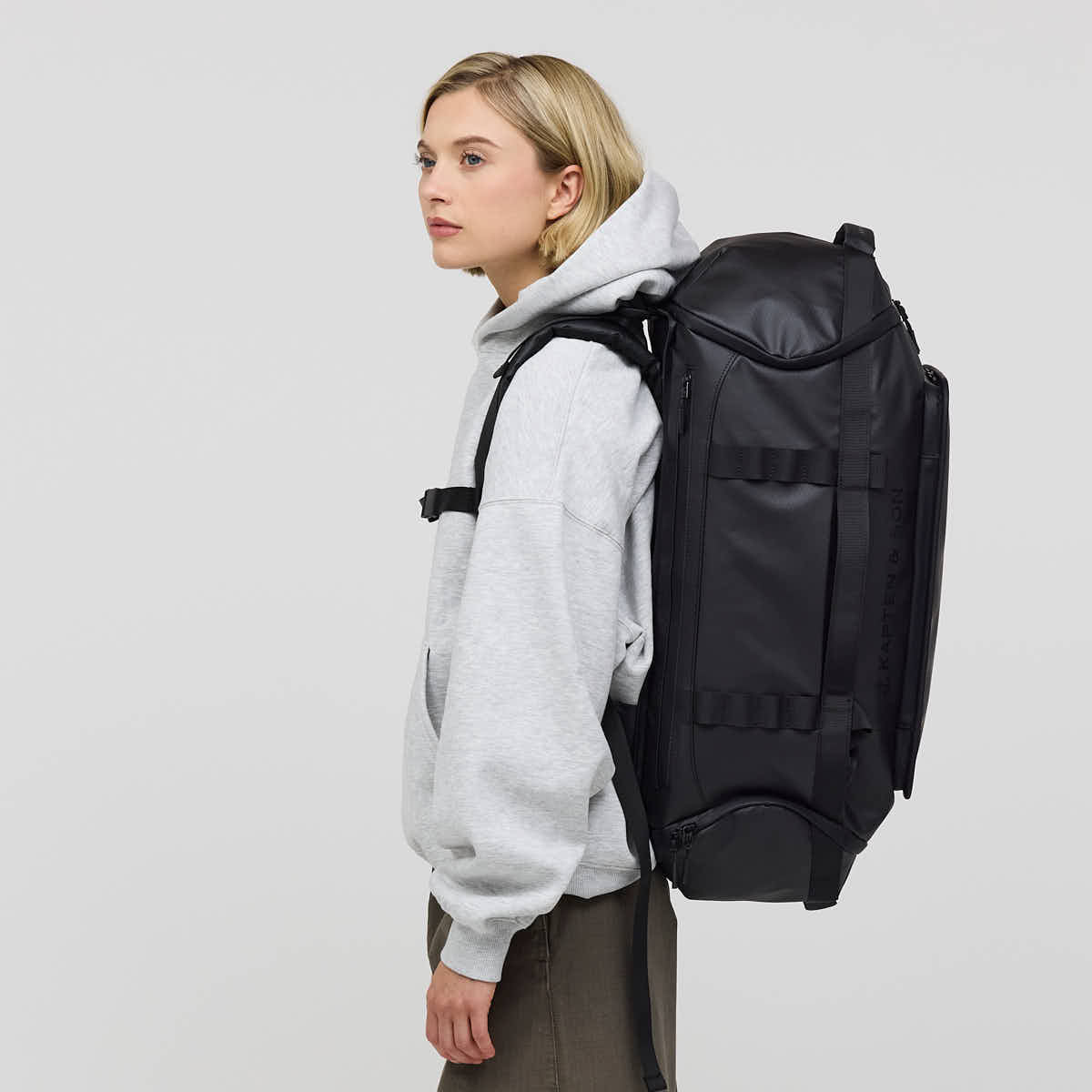 A woman wearing a light gray hoodie and dark pants is shown from the side, carrying a large black backpack on her back against a plain light gray studio background.