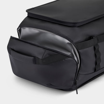 A close-up of a black waterproof backpack with its front zippered pocket open, showing a glossy black interior lining (waterproof) and a side carry handle, on a light gray background.