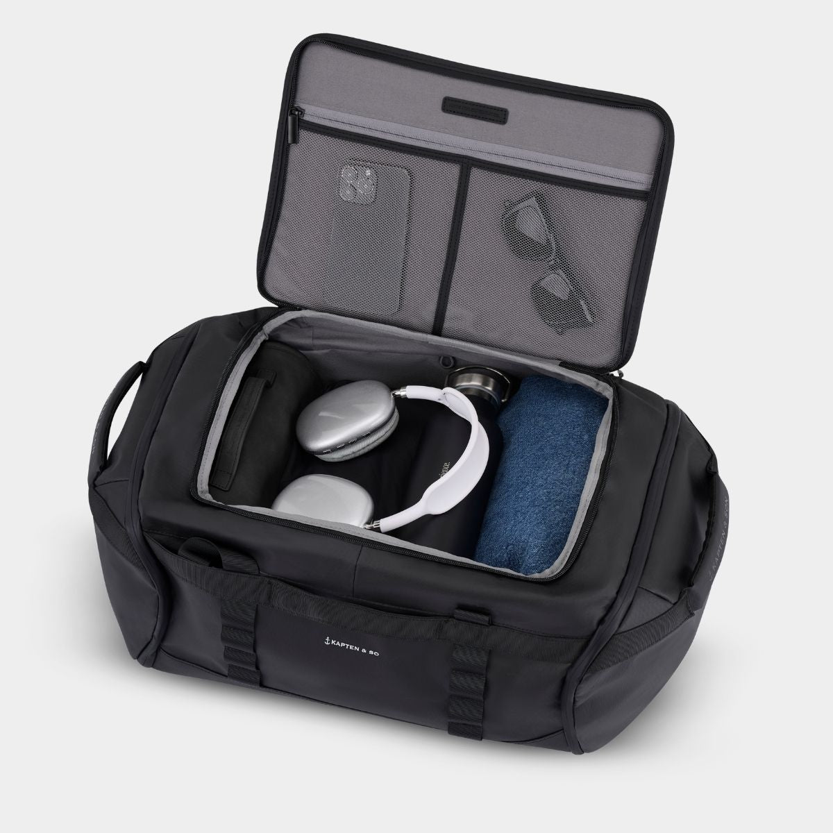 An open black duffel bag with gray interior lining, shown on a light gray background. The bag is packed with travel accessories, including white over-ear headphones, a jeans, sunglasses, and a smartphone in the organizer compartments.