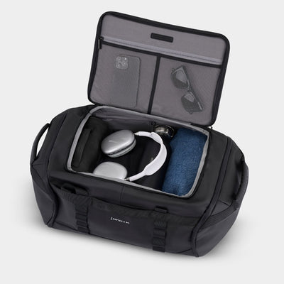 An open black duffel bag with gray interior lining, shown on a light gray background. The bag is packed with travel accessories, including white over-ear headphones, a jeans, sunglasses, and a smartphone in the organizer compartments.