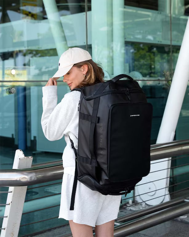 A woman wearing a white sweatshirt, shorts, and a white cap is shown from the back, carrying a large black travel backpack with multiple compartments and straps in an outdoor modern setting with glass buildings.