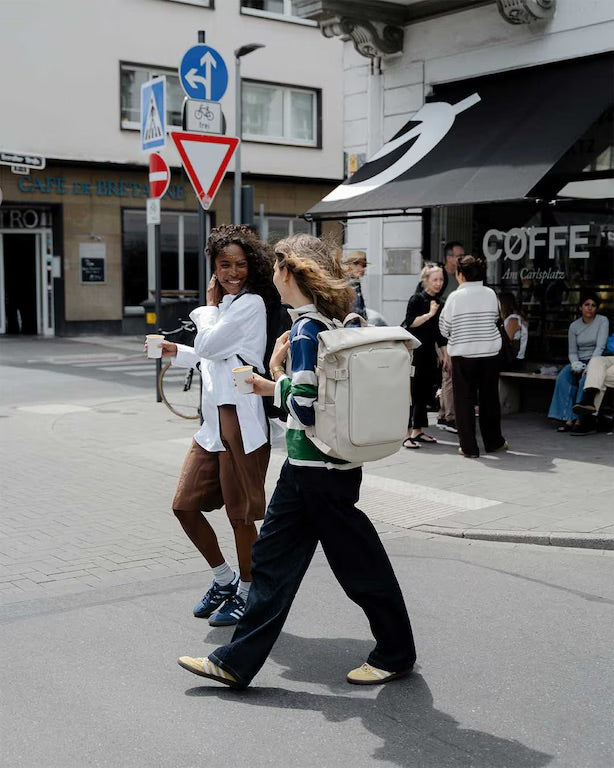 Two women walking on a city street, one wearing a white shirt and brown pants, the other in a green and white top with dark pants and a large off-white backpack. A coffee shop and street signs are visible in the background.