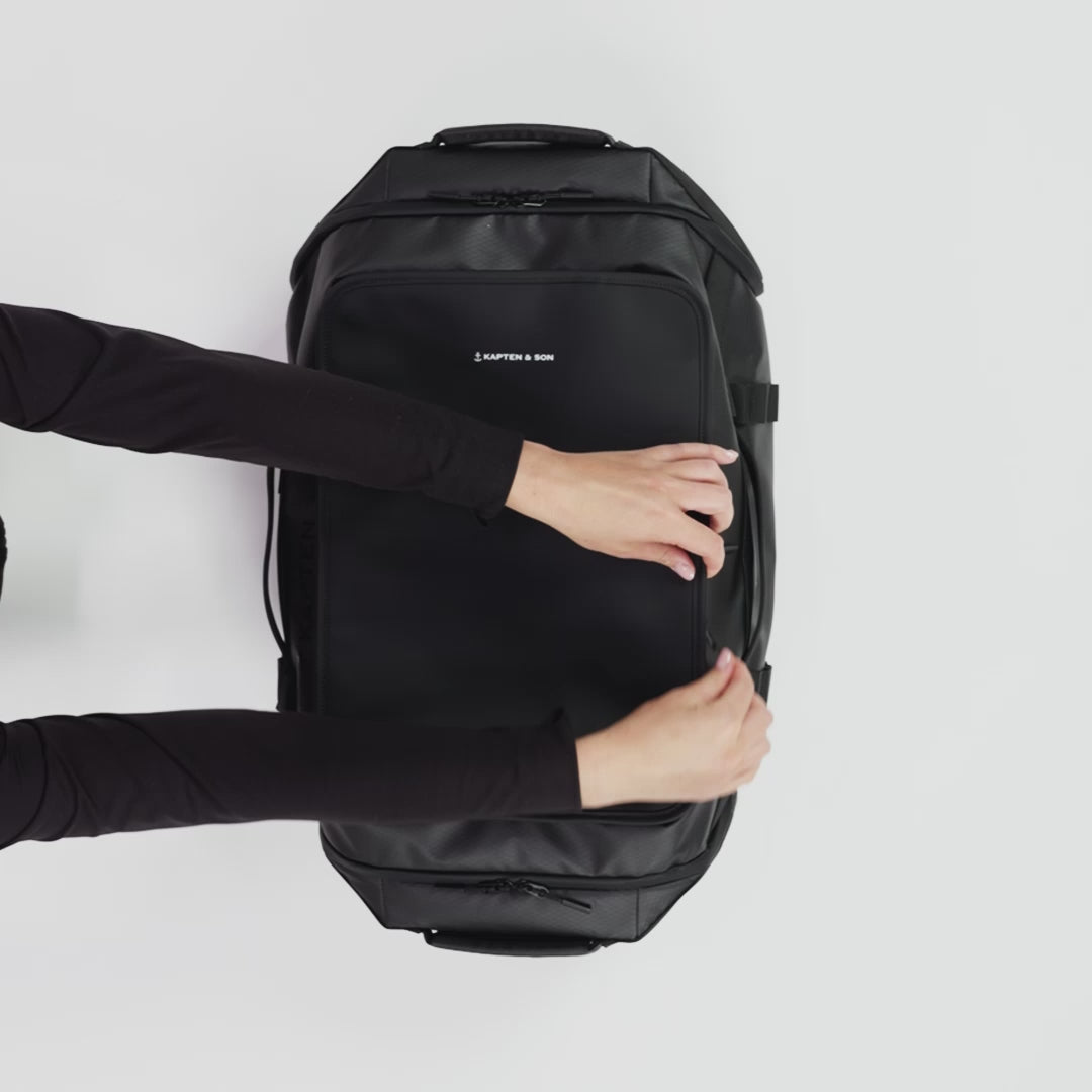 Video of black duffle bag opened from the top packed with a towel, toiletries, clothes, accessories, camera and headphones. Side pockets are opened to stores shoes and the backside for a laptop compartment.